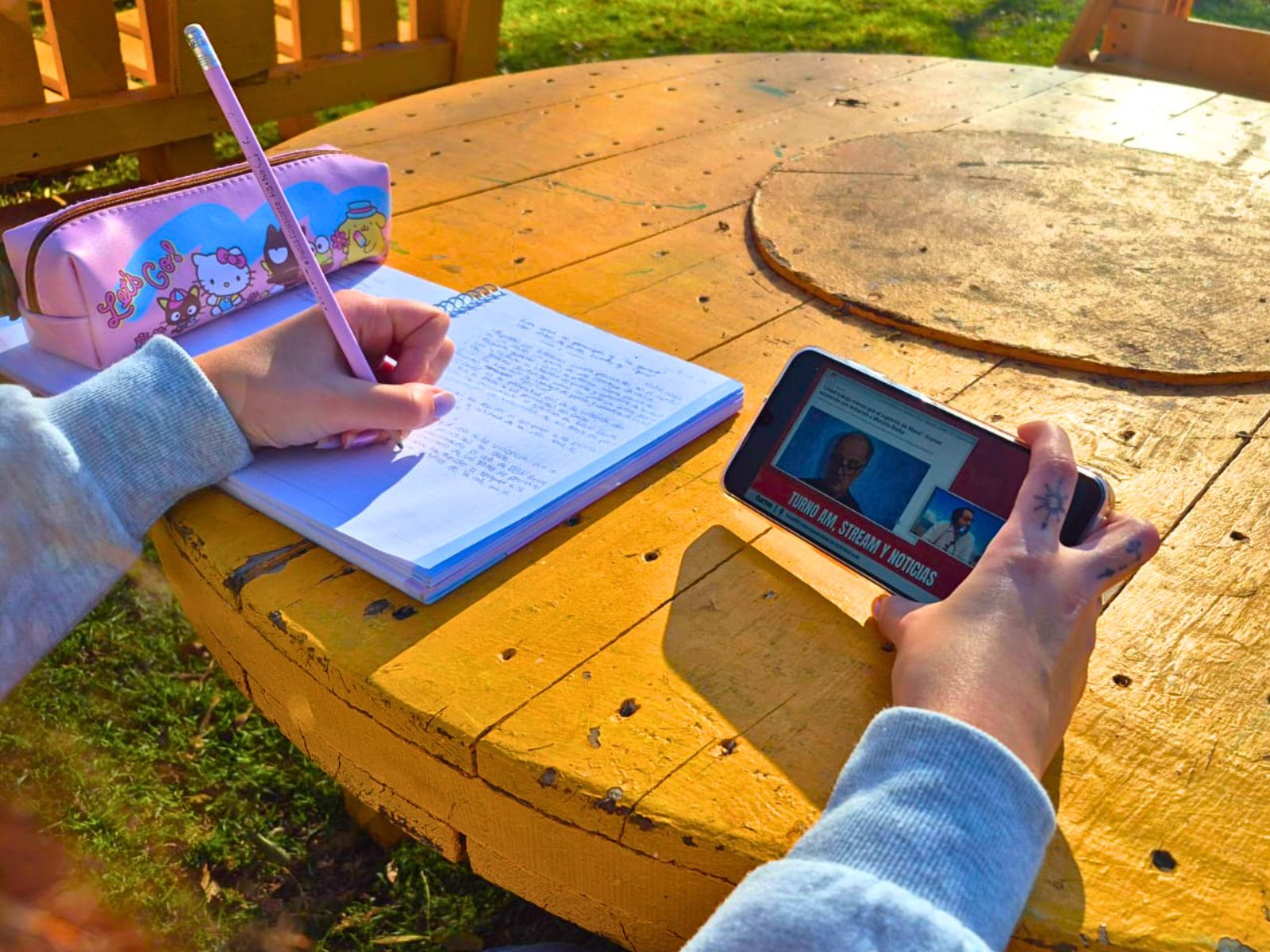 Manos de una persona joven que escribe en un cuaderno mientras sostiene un teléfono con contenido de noticias en redes sociales